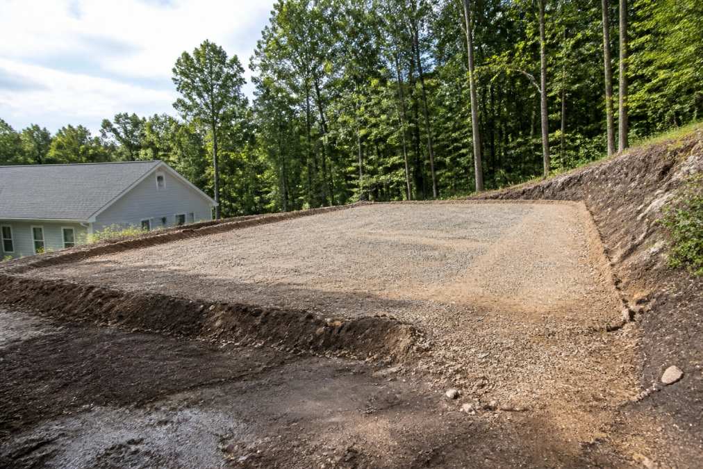 Residential property with uneven terrain and a cleared foundation area prepared for structure installation, showing graded soil and compacted base ready for garage placement
