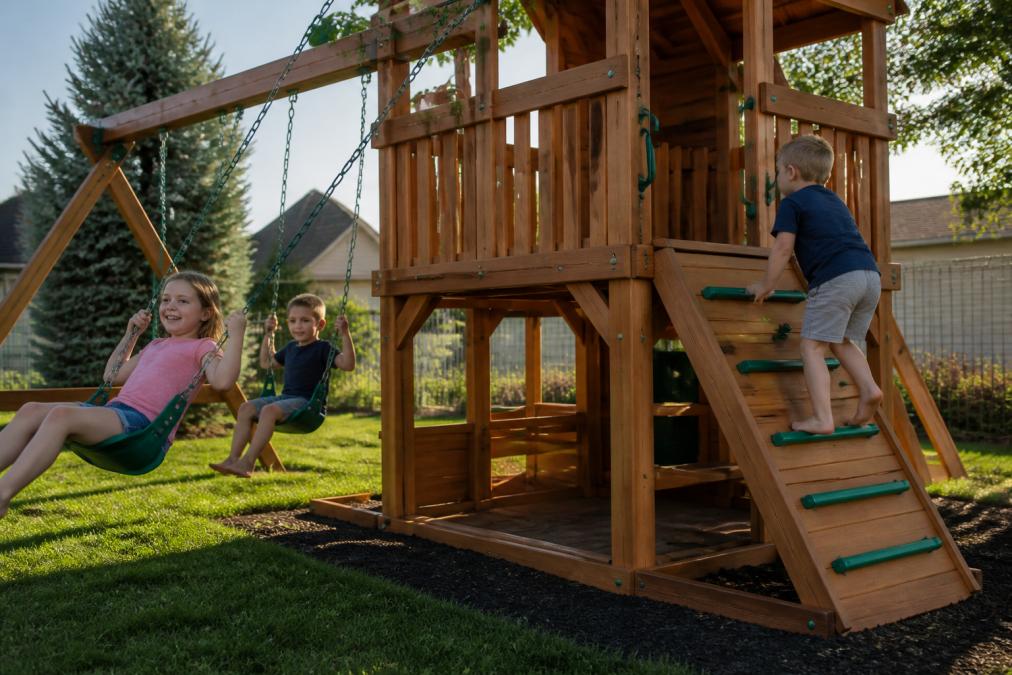 Young children actively playing on completed Amish-built wooden playset in suburban backyard, showing rubber-coated swing chains in motion, child climbing non-slip ladder rungs, and safety railings visible in afternoon sunlight