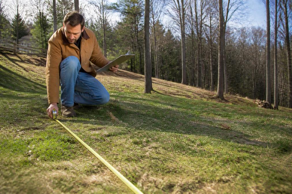 Homeowner in work clothes measuring their yard with a metal tape measure, examining the slope and terrain of their property with a clipboard in hand, natural daylight