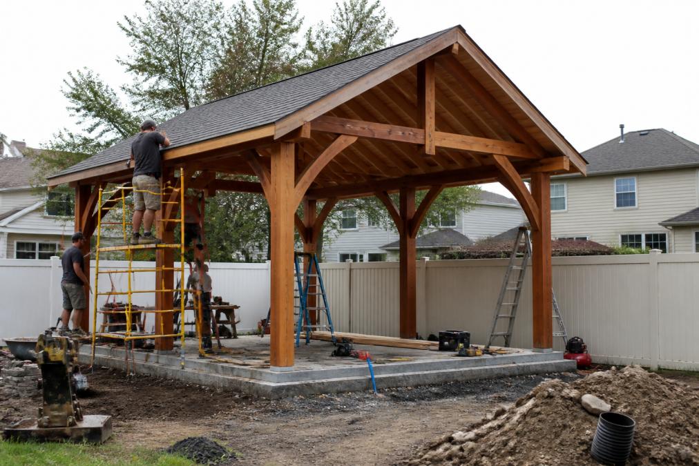 Professional construction crew installing a finished pavilion structure on a residential backyard, showing foundation work and structural assembly in progress with proper equipment and scaffolding