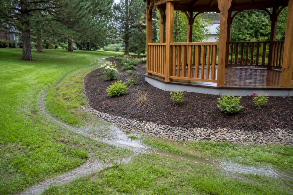 Sloped yard showing proper drainage grading away from a gazebo foundation with visible water flow patterns and landscaping that directs moisture toward the perimeter