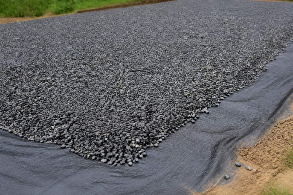 Close-up of compacted gravel base with dark landscape fabric visible underneath, showing proper gravel pad preparation with crushed stone evenly distributed, natural daylight