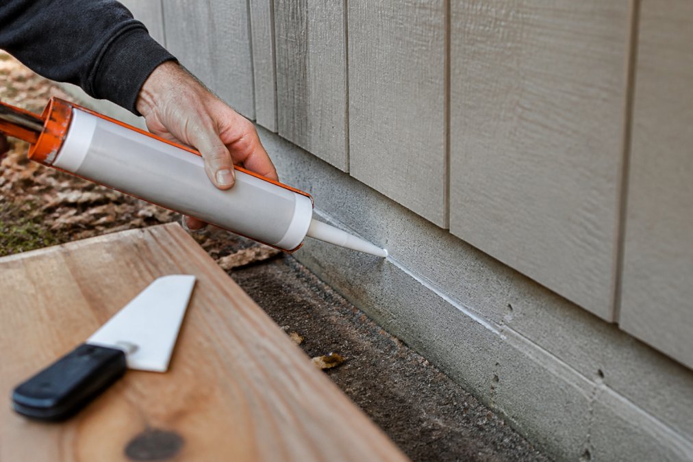 Close-up of a hand applying exterior caulk to a gap between shed wooden siding and concrete foundation, with a caulk gun and smoothing tool visible on a workbench