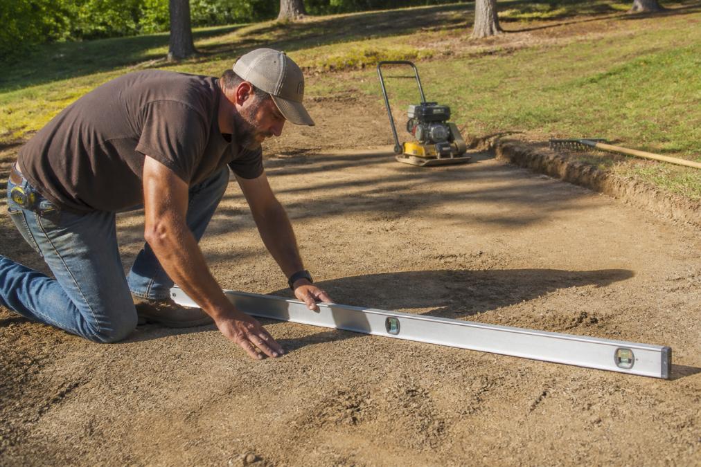 Homeowner using a long aluminum level to prepare shed foundation by checking ground evenness on a sloped yard