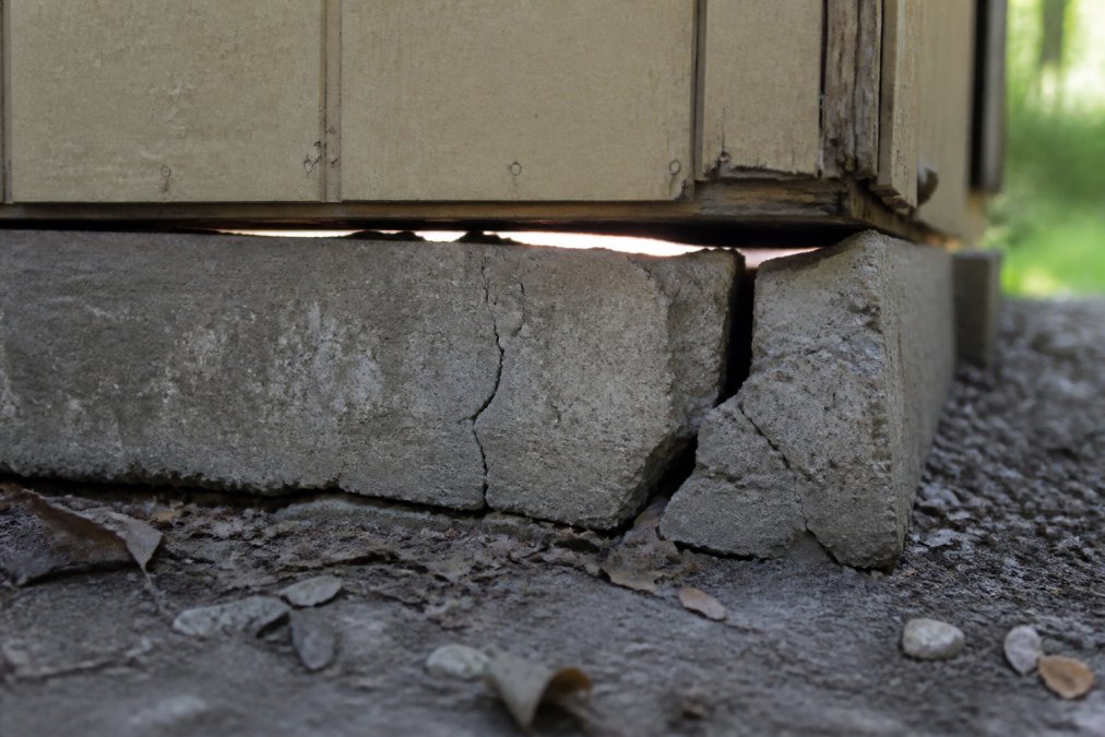 Close-up of concrete foundation showing visible cracks, settling, and separation along the edges where the shed base meets the ground, with daylight visible through gaps
