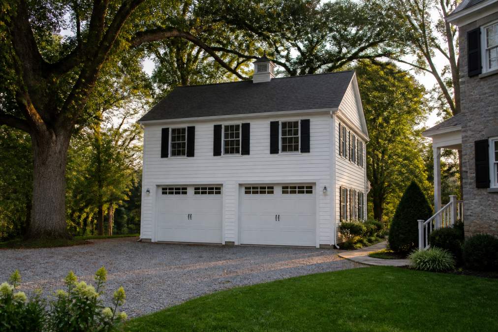 A two story detached garage with white siding and dark shutters on a residential West Chester PA property, gravel driveway, mature oak trees, late afternoon light